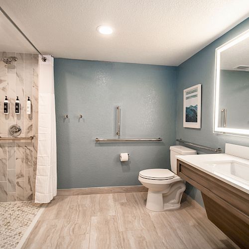 A modern bathroom with a walk-in shower, wall-mounted toiletry dispensers, towels, toilet, sink with large mirror, and safety grab bars.