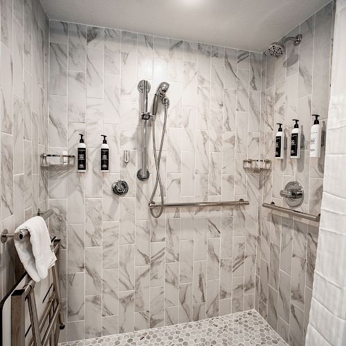 A modern shower with marble tiles, multiple showerheads, soap dispensers, a towel rack, and grab bars, enclosed by a shower curtain and tiles.