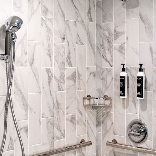 A marble-tiled shower with a handheld showerhead, soap dispensers, grab bars, and a small shelf is shown in this image.