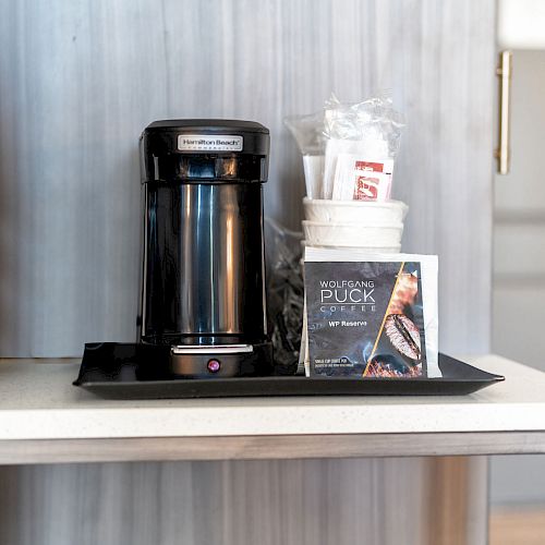 A coffee station with a coffee maker, disposable cups, stirrers, sugar packets, and a Wolfgang Puck coffee pod on a black tray.