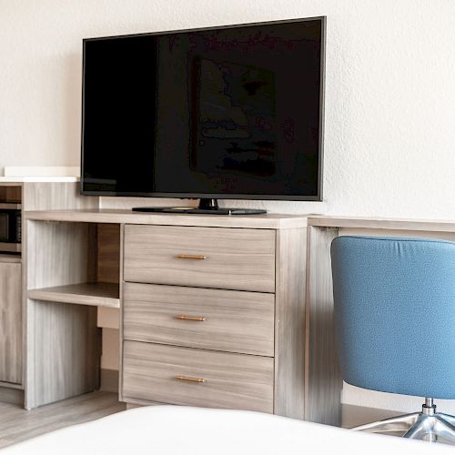 A modern hotel room setup with a large TV, wooden cabinets, a microwave, and a blue chair at a desk.