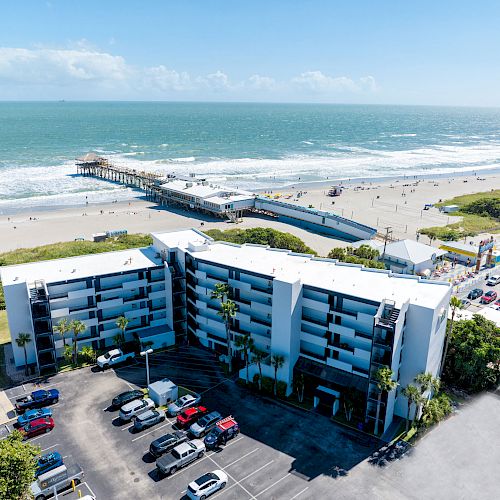 Aerial view of a beachfront building with a pier extending into the ocean, cars in the parking lot, and people on the beach.