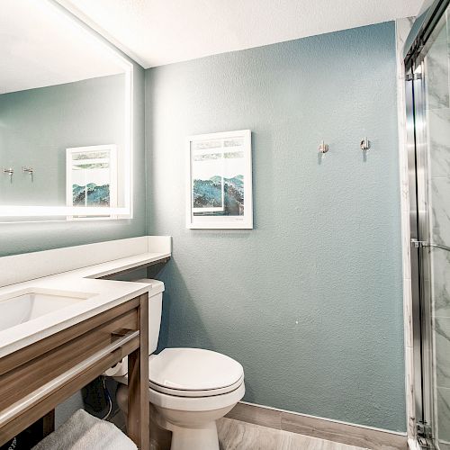 This is a modern bathroom with a vanity, sink, mirror, toilet, and a glass-enclosed shower. The walls are a light teal color with a framed picture.