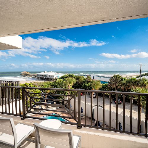 A balcony overlooking a scenic beach view with chairs and a table, clear blue sky, and greenery in the foreground, ending the sentence.