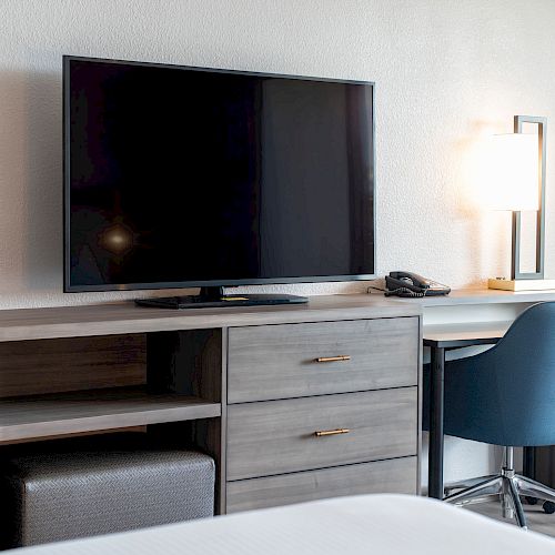 A hotel room setup with a flat-screen TV, a desk with a lamp, phone, and books, and a blue chair. The desk has a modern wooden design.