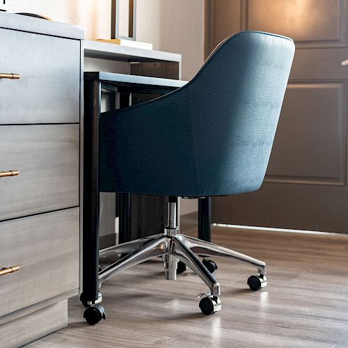 A modern home office setup featuring a blue swivel chair on casters, a desk, and a chest of drawers, with a door in the background.