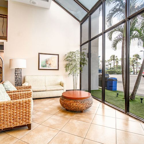 A bright room with large windows features two wicker chairs, a beige sofa, a round wooden table, a lamp, and a painting, overlooking a parking lot.