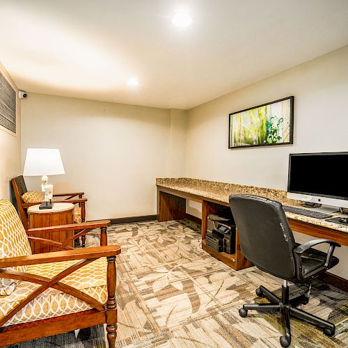 A small office room featuring a desk with a computer, a rolling chair, two cushioned chairs, a lamp, and a wall picture.