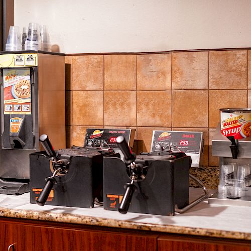 A breakfast station setup with a waffle maker, batter dispensers, syrup, and plastic cups on a countertop by a tiled wall, ready for use.