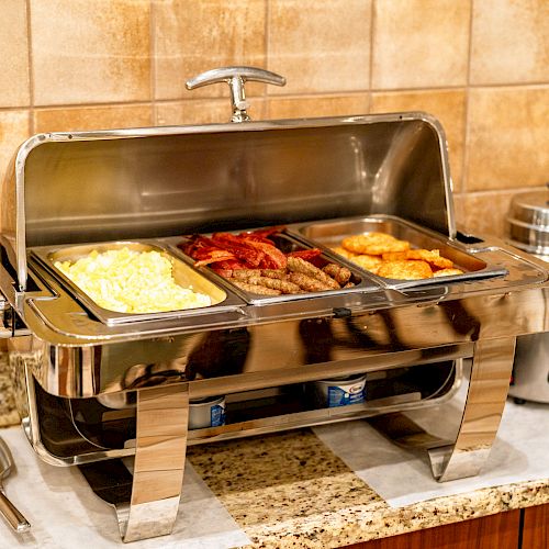 The image shows a buffet setup with scrambled eggs, bacon, sausages, and fried potatoes in a stainless steel chafing dish on a countertop.