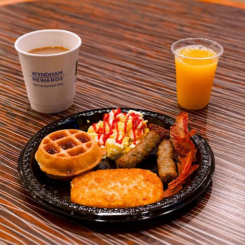 A breakfast plate with a waffle, hash brown, scrambled eggs with ketchup, bacon, and sausage is seen alongside a cup of coffee and a glass of orange juice.