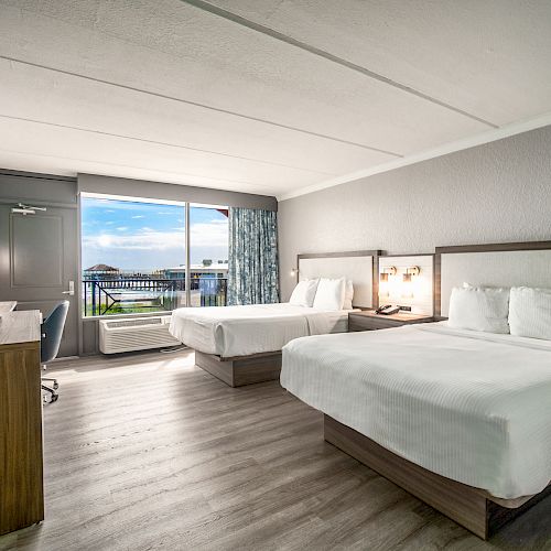 This image shows a bright hotel room with two beds, a TV, a desk, and a sliding glass door leading to a balcony.