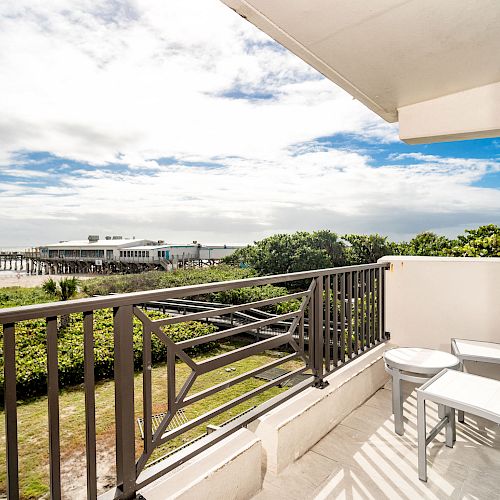 A balcony with a table and two chairs overlooks a scenic pier, beach, and ocean view under a partly cloudy sky, surrounded by lush greenery.