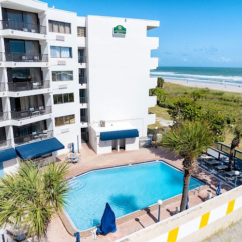 This image shows a beachfront hotel with a swimming pool, surrounded by palm trees. The building has multiple balconies with ocean views.