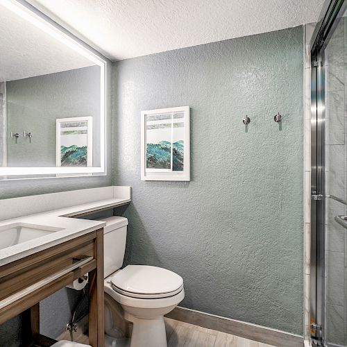 A modern bathroom with a sink, toilet, and glass shower. The walls are gray, featuring two framed pictures, and the space has a large mirror and shelf.