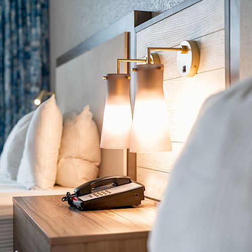 A tidy hotel room features a bed with white pillows and sheets, an illuminated bedside lamp, and a telephone on the wooden bedside table.