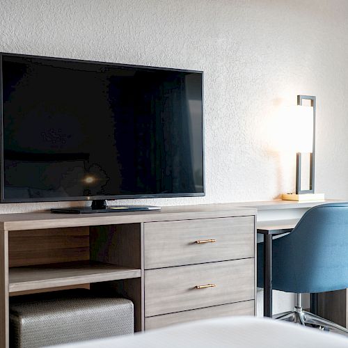 The image shows a modern hotel room setting with a large TV on a wooden cabinet, a desk with a lit lamp, and a blue chair next to it.