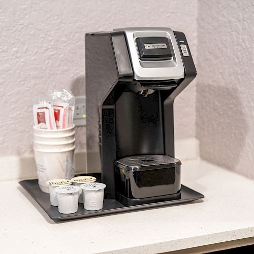 A single-serve coffee maker sits on a counter next to a stack of cups, creamer cups, and stirrers in packets, all organized on a tray.