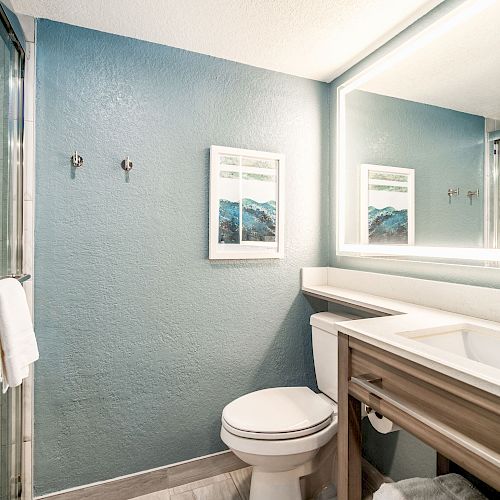 A modern bathroom with a glass shower, toilet, wooden vanity with sink, large mirror, and wall art. The walls are painted blue.