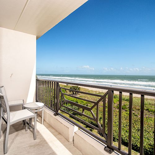 A balcony with a table and two chairs overlooks a scenic beach with clear skies and ocean waves, offering a tranquil and inviting view.