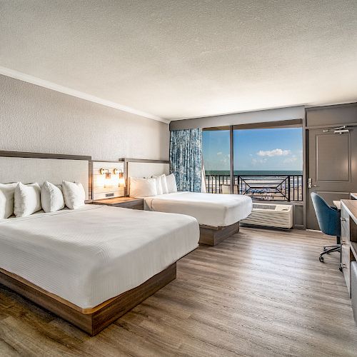 A hotel room with two large beds, a TV, a desk with a chair, and a window showing a balcony with an ocean view in the background.