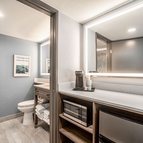 A modern bathroom setup with a toilet, vanity, illuminated mirror, and open shelving holding towels and a mini-fridge, completed with light fixtures.