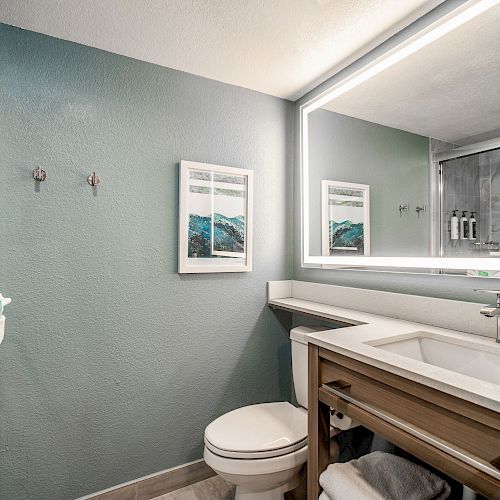 A modern bathroom with a large illuminated mirror, sink, toilet, and shower with glass doors. Two framed artworks are hung on the wall.