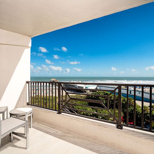 A balcony with chairs and a small table, overlooking a beautiful beach and ocean under a clear blue sky with scattered clouds.