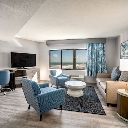 A modern living room with blue accents, featuring a sofa, armchairs, a coffee table, a TV on a desk, and a window with ocean views.