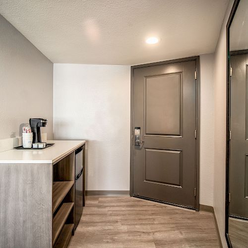 The image shows a modern entryway with a coffee station on a counter, a large mirror on the wall, light wood flooring, and a closed door.