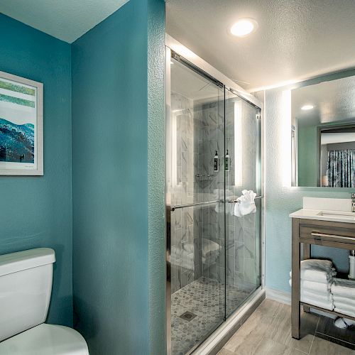 A modern bathroom with teal walls, a framed picture, a toilet, a glass-enclosed shower, and a vanity with a lighted mirror and storage shelves.