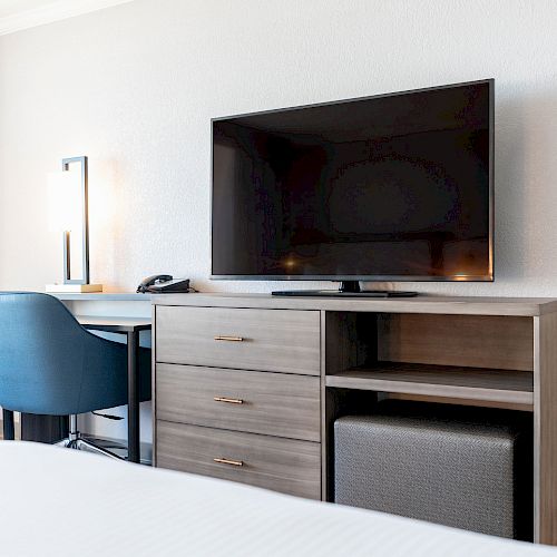 The image shows a modern hotel room setup with a flat-screen TV on a dresser, a desk with a chair, a lamp, and a phone next to a bed.