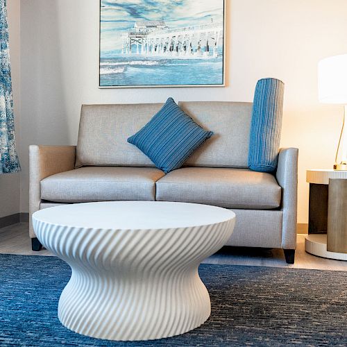 A living room with a beige sofa, blue cushions, modern white table, blue rug, and a lit lamp on a round side table.