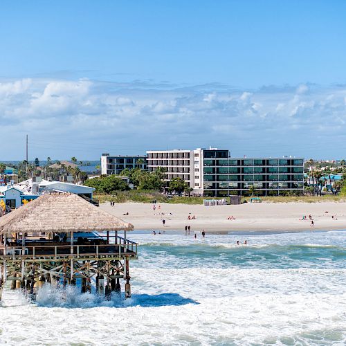 A beach scene with a pier extending into the ocean, waves crashing, people on the beach, and buildings in the background under a partly cloudy sky.