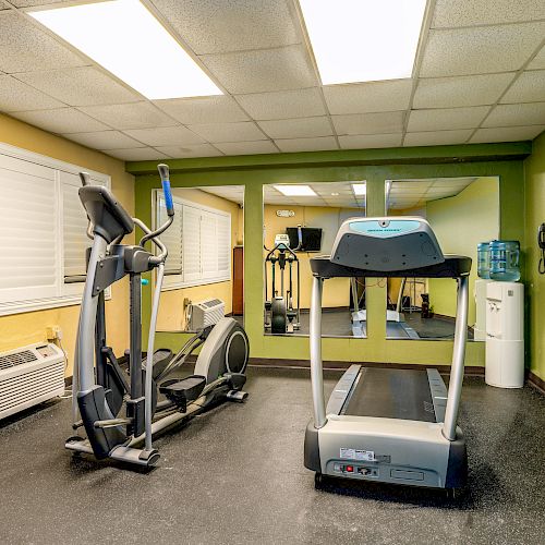 A small gym with an elliptical machine, treadmill, wall mirror, air conditioning unit, and water cooler in a brightly lit room with green walls.