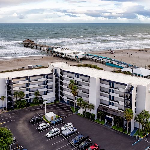 The image features a beachfront hotel with a parking lot and a pier extending into the ocean under a partly cloudy sky.