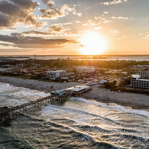 An aerial view of a coastal town during sunset, with a long pier extending into the ocean and buildings lining the shoreline.