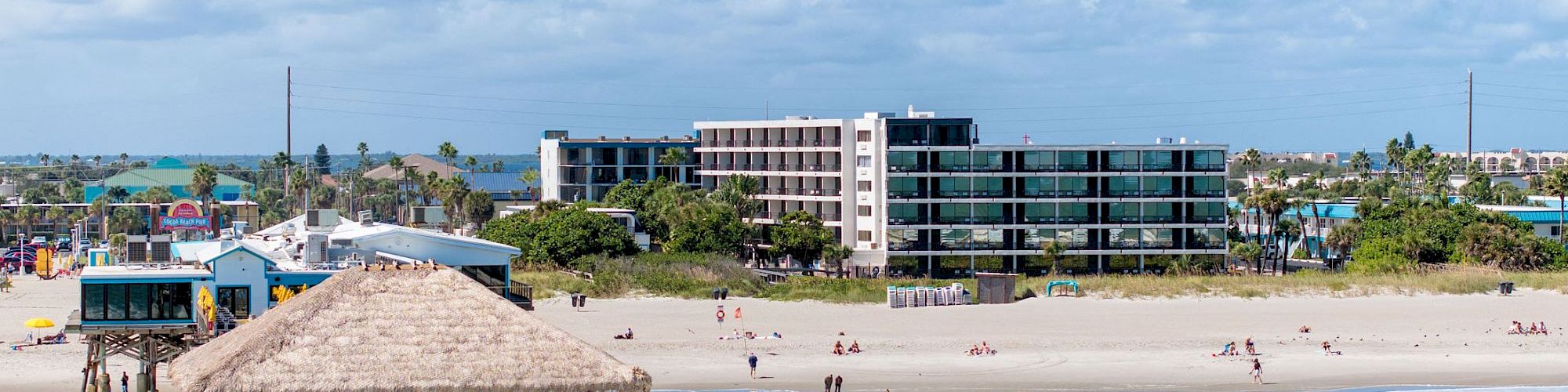 A beachfront scene with a pier and thatched roof hut over the water, people on the beach, and several buildings in the background against a blue sky.