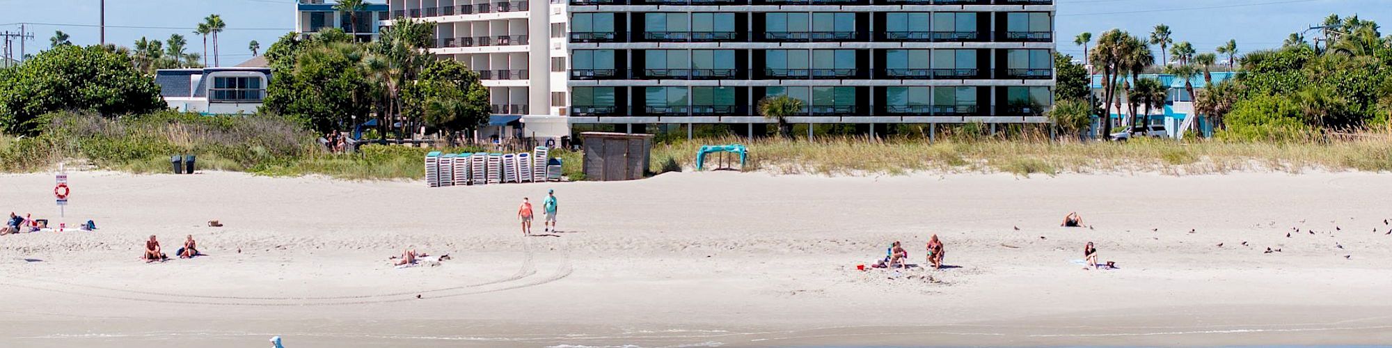 A beachside scene with a few people scattered on the sand. In the background are buildings including a multi-story hotel or apartment complex.