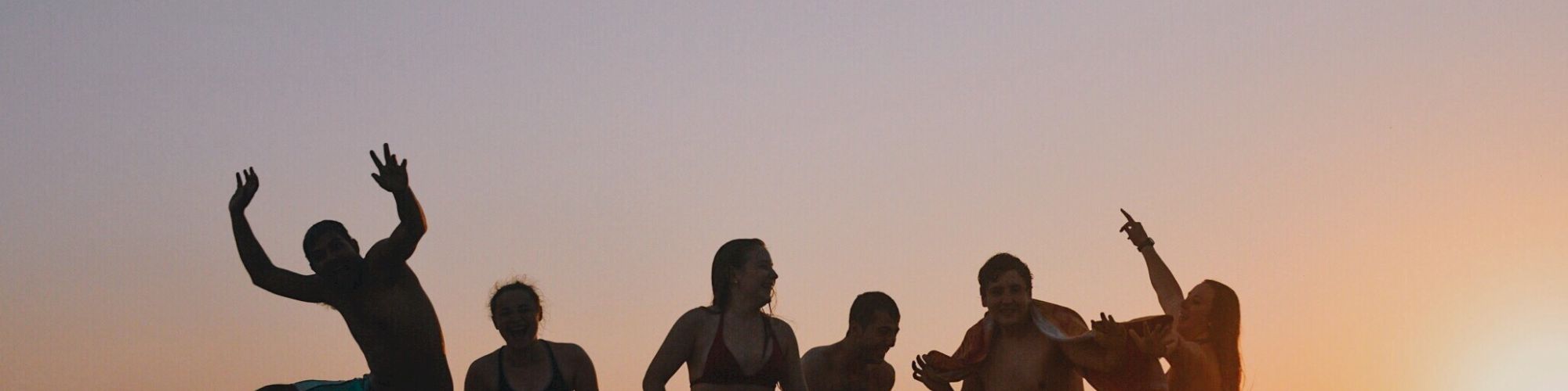 A group of people are having fun on a beach at sunset, with some jumping and posing playfully against the scenic background.