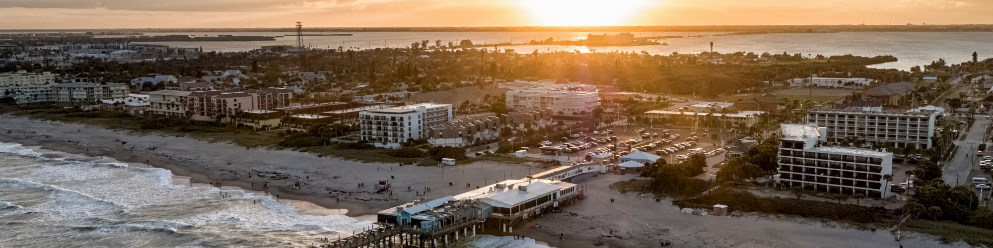 Aerial view of a coastal city during sunset, with a pier extending into the ocean and waves crashing onto the shore.