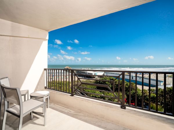 A balcony with two chairs and a table overlooks a sunny beach and ocean under a clear blue sky with white clouds.