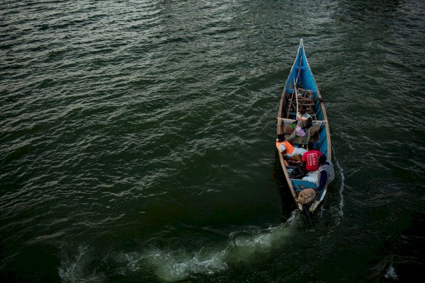 A small boat with three people, one wearing red, is moving through green waters, leaving a trail behind them, under a cloudy sky.