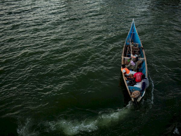 A small boat with three people, one wearing red, is moving through green waters, leaving a trail behind them, under a cloudy sky.