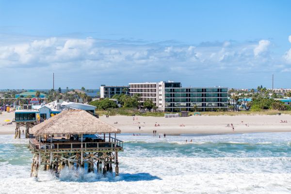 A beach scene with a pier over the water, people on the sand, and buildings in the background under a clear blue sky.
