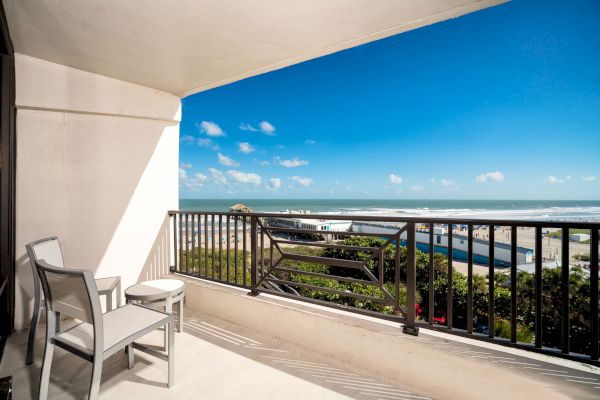 A balcony with two chairs and a table overlooks the ocean under a clear blue sky with scattered clouds in the distance.