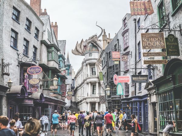 A bustling street scene with people walking, lined with shops, featuring a large dragon on a rooftop, evoking a magical atmosphere.