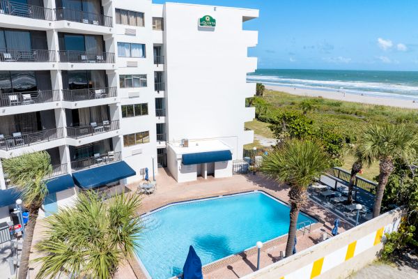 The image shows a hotel building with a swimming pool, palm trees, and a beach view on a sunny day.