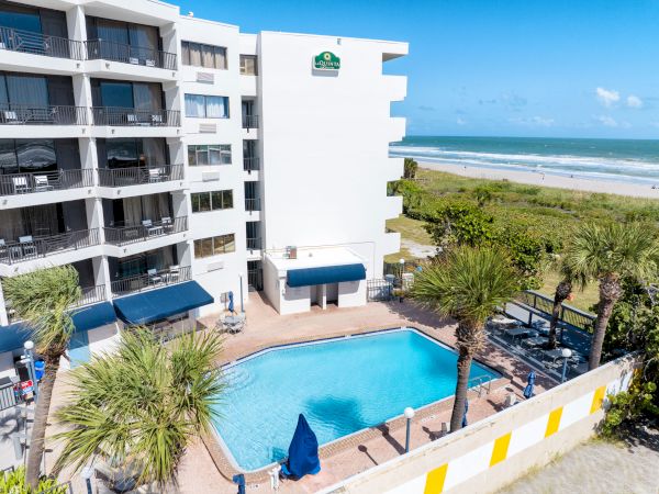 The image shows a hotel building with a swimming pool, palm trees, and a beach view on a sunny day.
