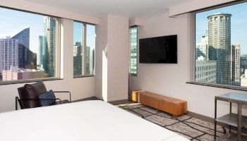The image shows a modern hotel room with large windows offering city views, a wall-mounted TV, a chair, a low table, and a bed.